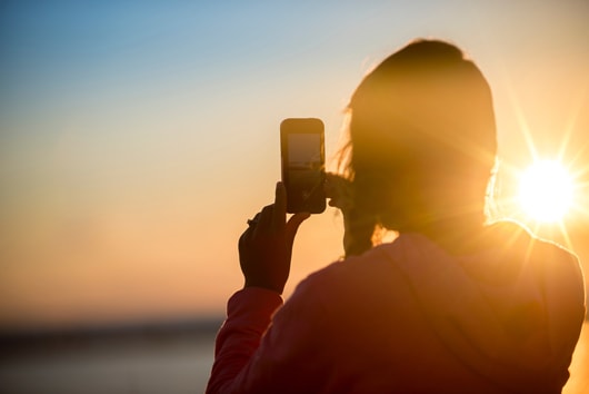 The silhouette of a person from behind, taking a picture of the setting sun in the background.